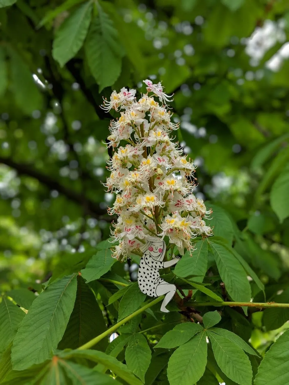 Tiny paper girl seated in horse-chestnut leaves holding a white flower spike like a giant bouquet