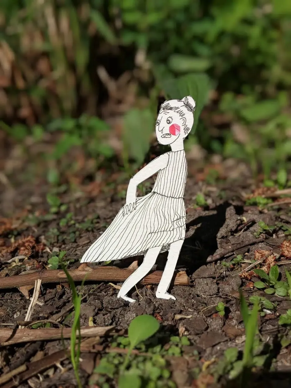 Cut-out woman in a striped dress, hand on hip, standing on a twig in dark soil