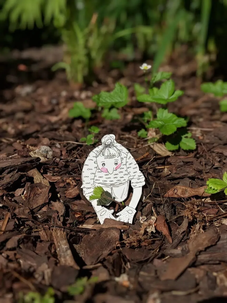 Round little figure in a looped-pattern jumper crouching on bark mulch, clutching a real green leaf