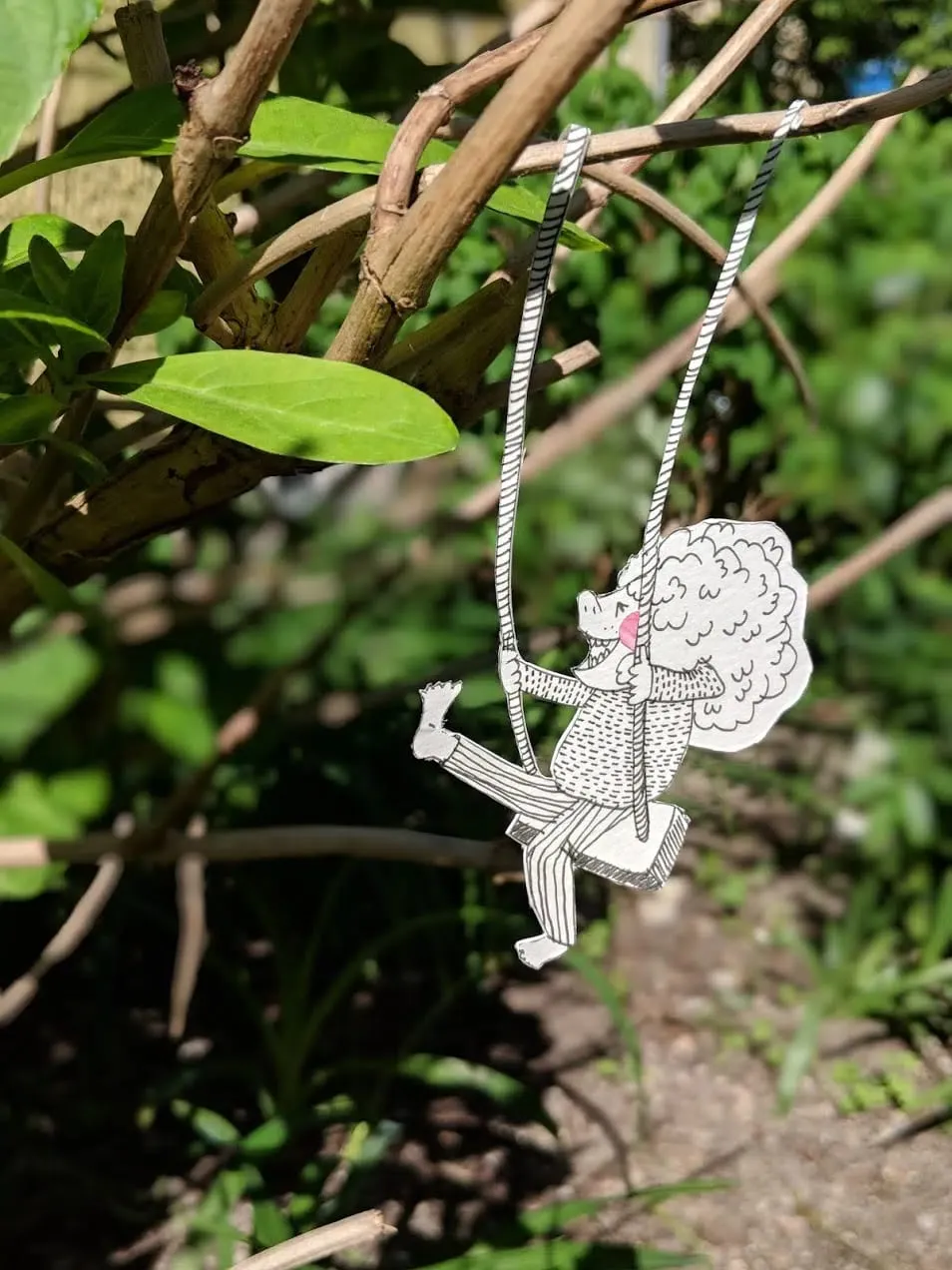 Curly-haired cut-out figure swinging on a paper swing hung from a real garden branch