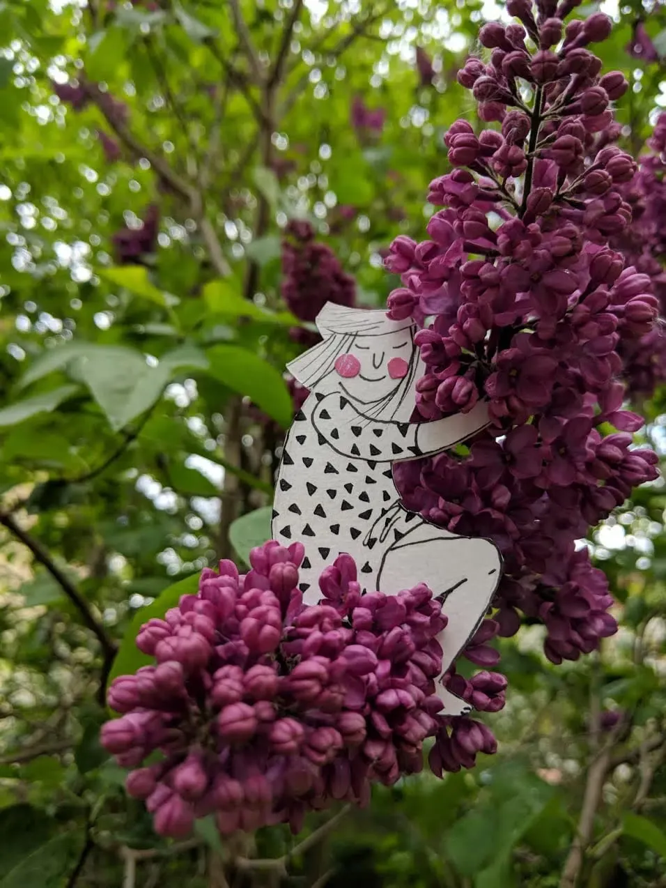 Long-haired cut-out girl in a triangle-print dress hugging a heavy purple lilac panicle