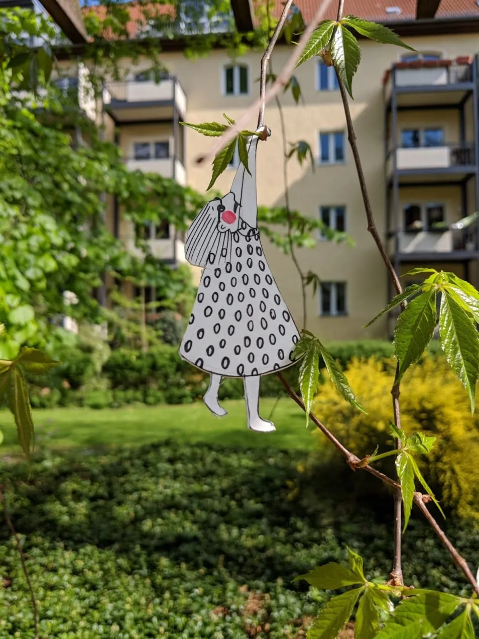 Cut-out girl in a spotted dress dangling one-armed from a thin branch above a green courtyard
