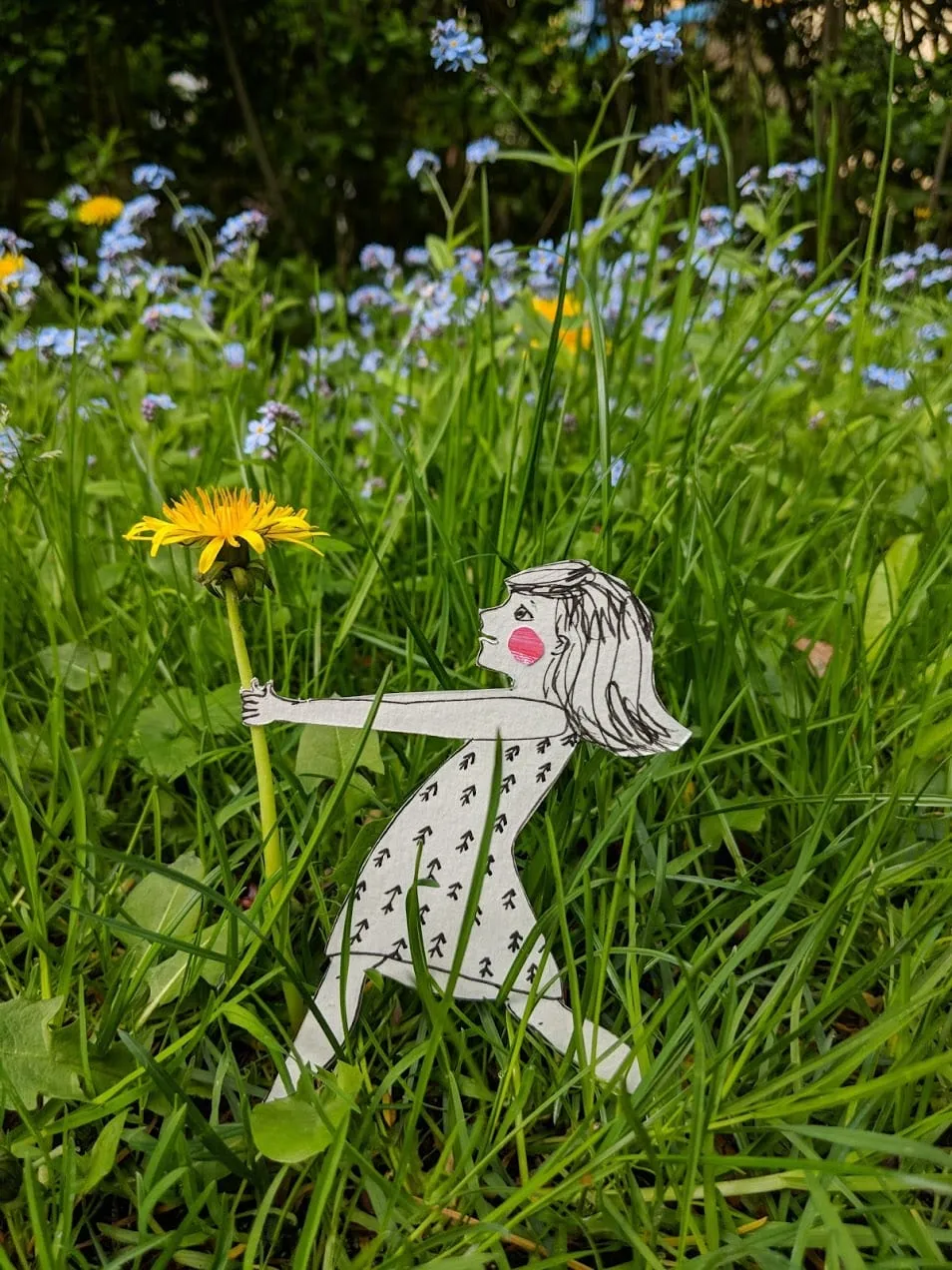 Girl in an arrow-print dress reaching to grasp a dandelion stem amid grass and blue forget-me-nots