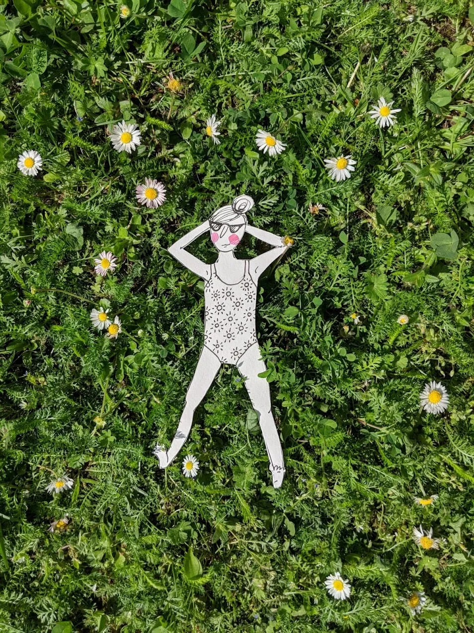 Cut-out sunbather in a patterned swimsuit and sunglasses lying flat in grass scattered with daisies