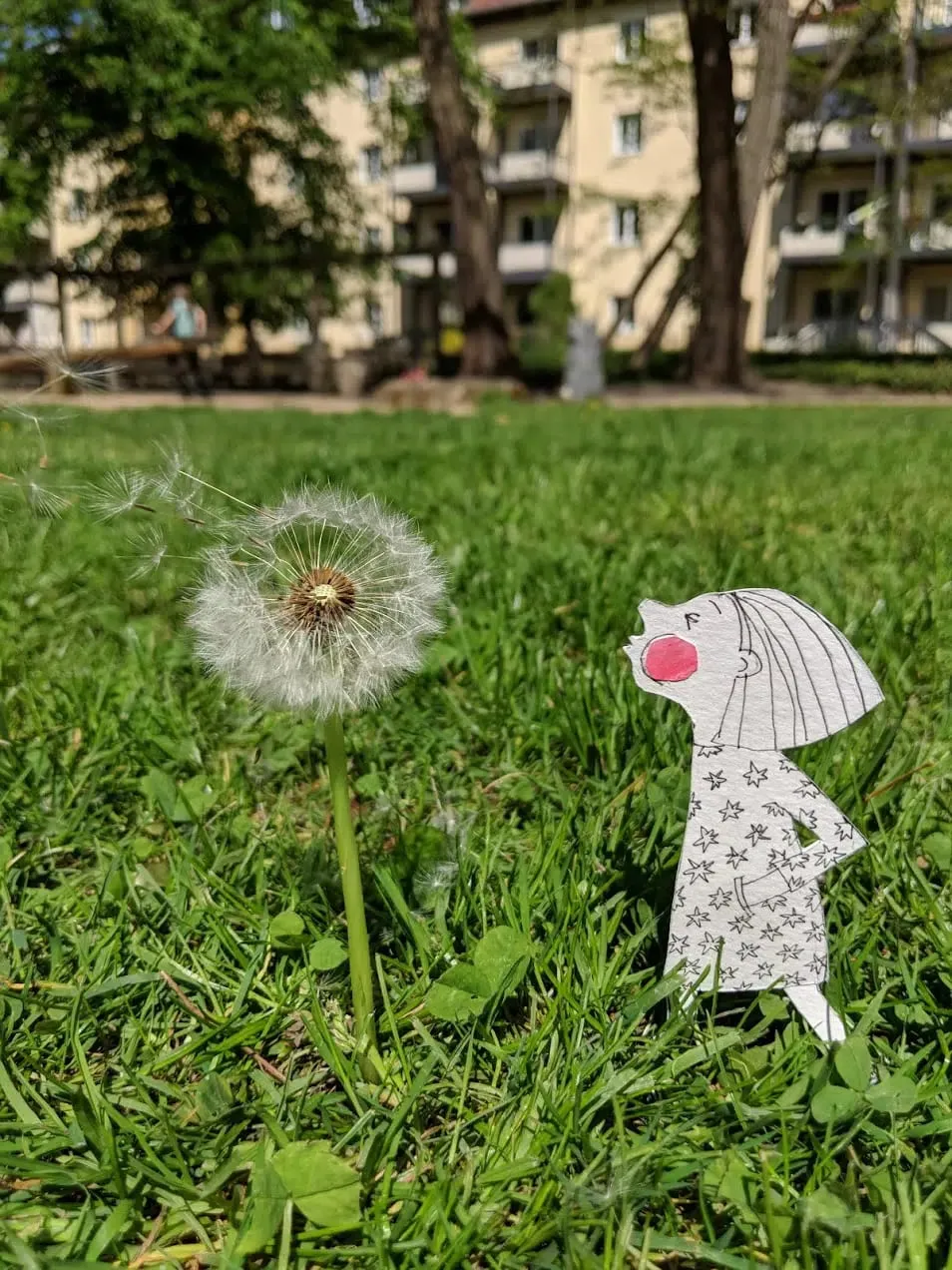 Girl in a star-print dress blowing a dandelion clock, seeds drifting across a sunlit park lawn