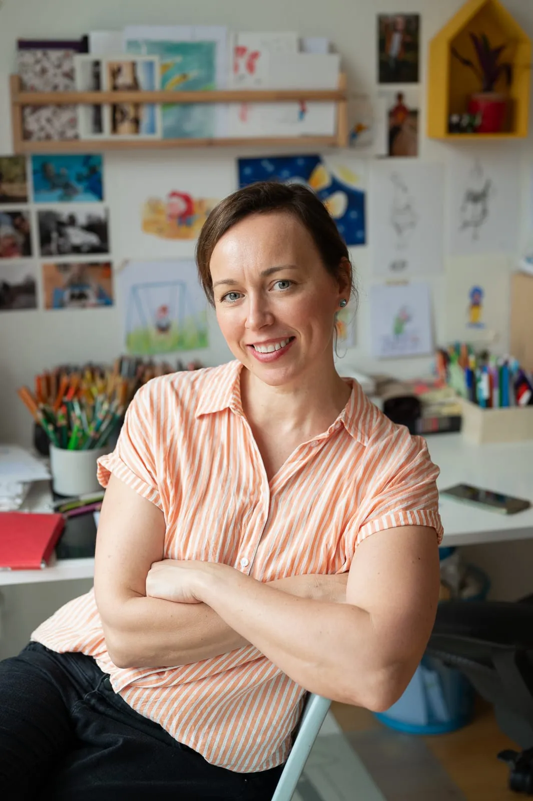 Tiiu Kitsik in her studio, arms folded and smiling, in front of a wall pinned with drawings, photographs and jars of coloured pencils