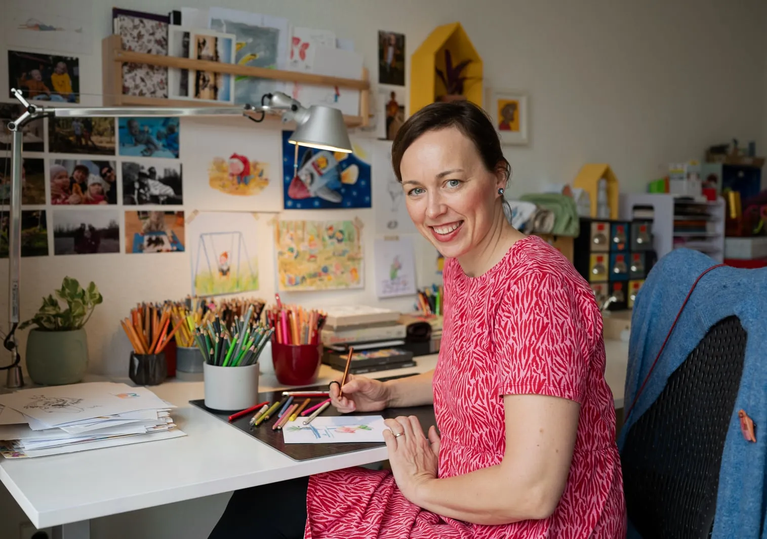 Tiiu Kitsik at her drawing table in a red patterned dress, colouring a sketch, her own illustrations covering the wall behind her